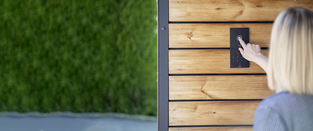 Woman pressing the bell of a Loxone Intercom in anthracite on timber cladding, shown with NFC Code Touch, sold separately